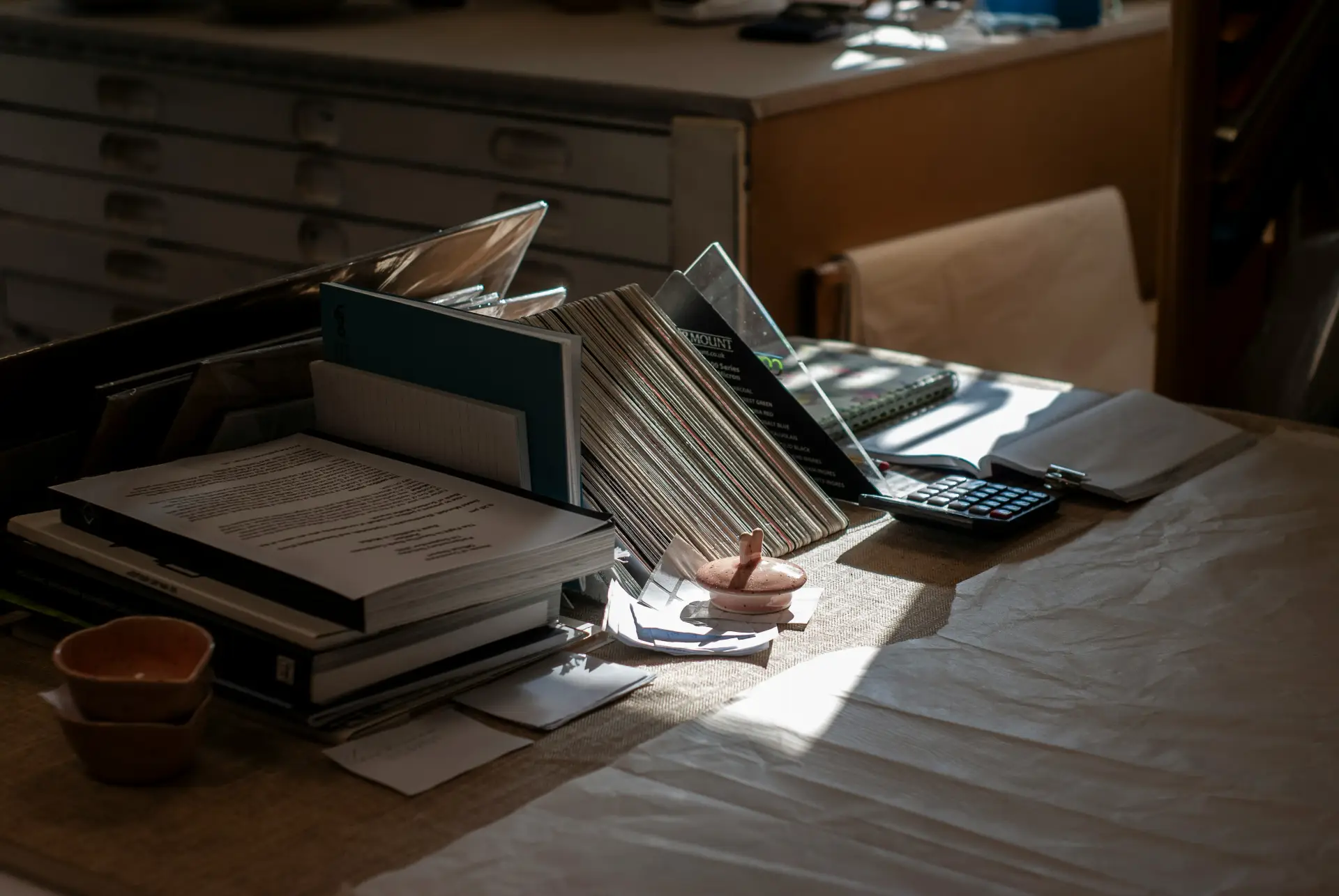 A desk in warm sunlight with stacked papers, a ledger and a calculator